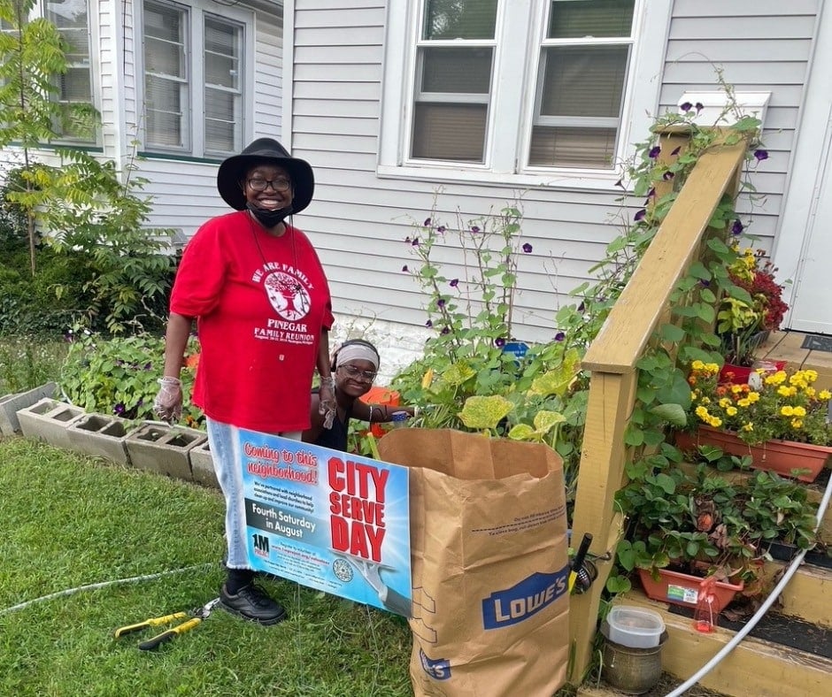A woman in a red t-shirt and black hat and a young person kneel beside a flower and vegetable garden in front of a house, holding a "City Serve Day" banner next to a Lowe's yard-waste bag.